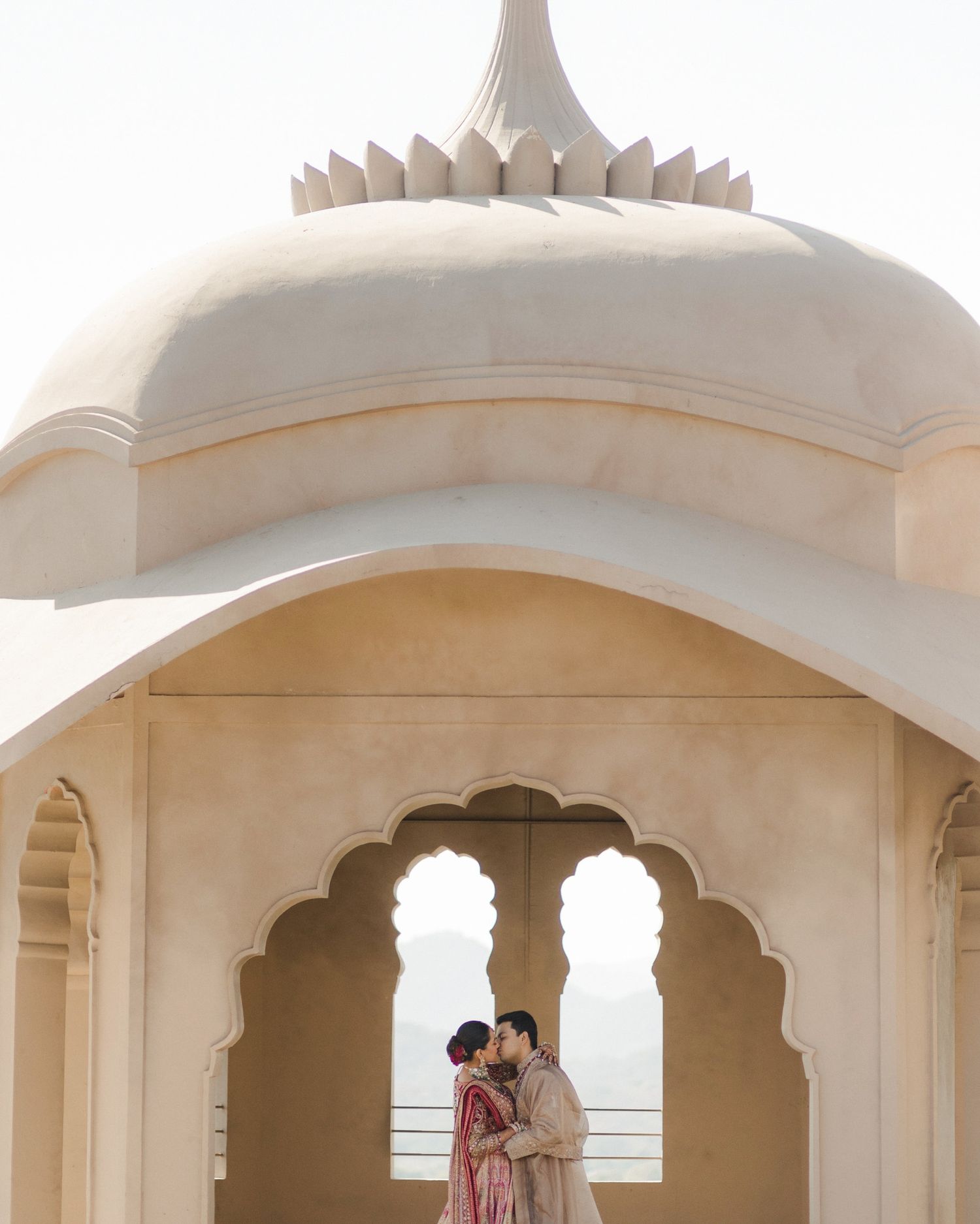 Indian bride and groom sharing an intimate moment under ornate white arches of a palace at their wedding.