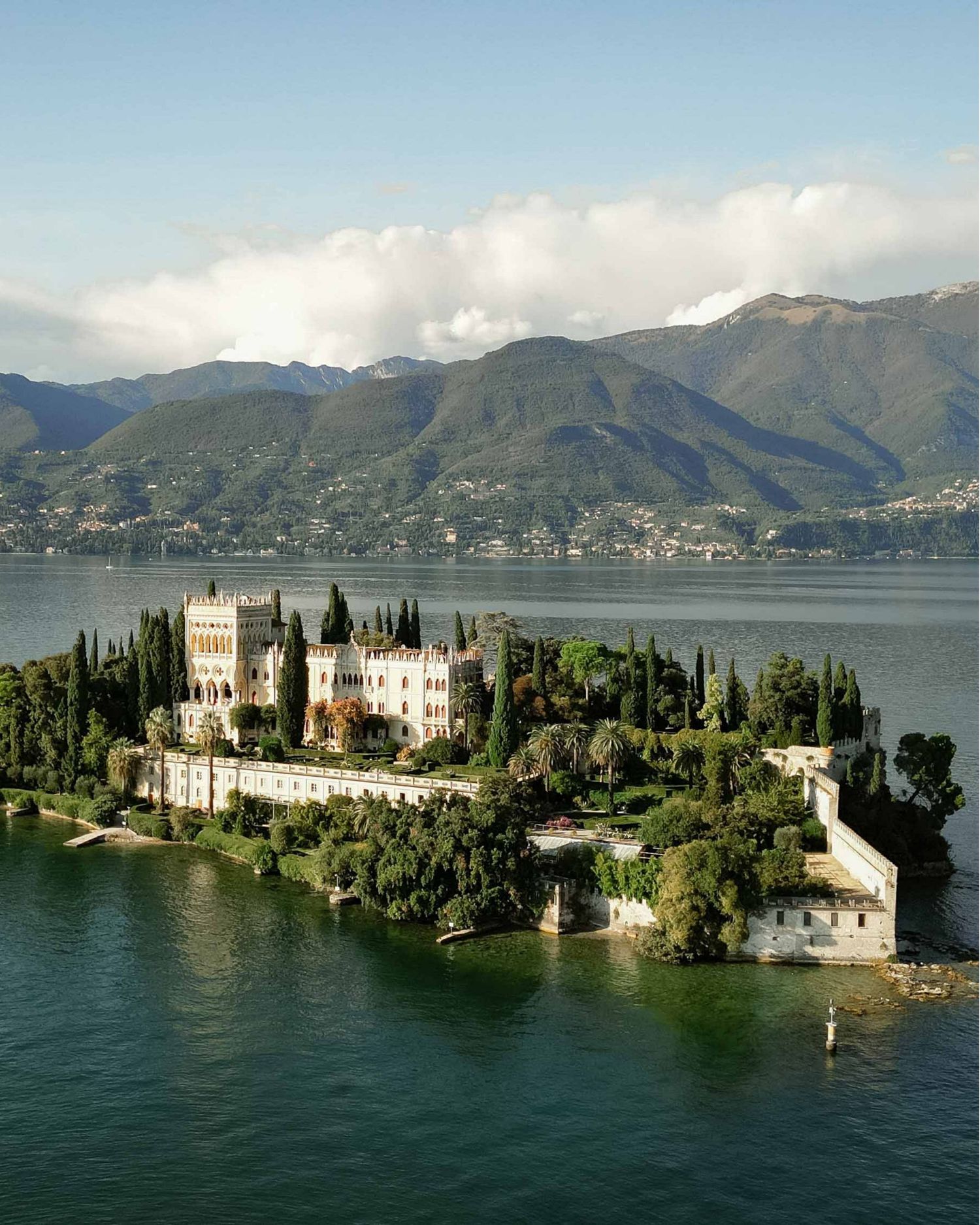 Aerial view of Isola del Garda castle surrounded by lush gardens on Lake Garda with mountain backdrop.