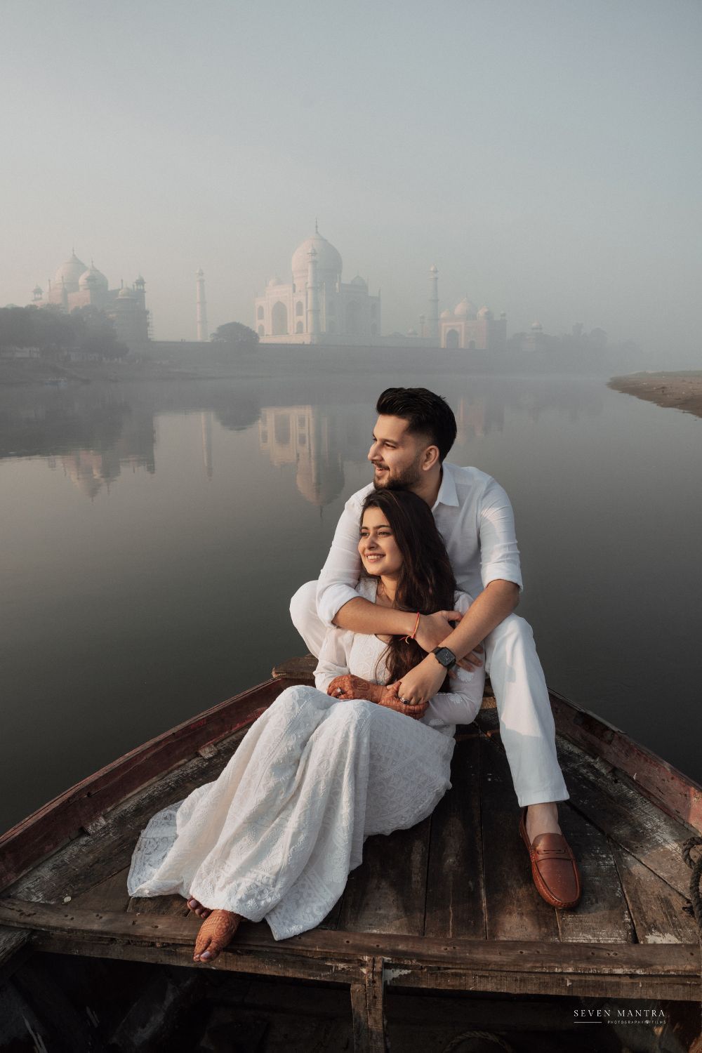 Couple embraces on a wooden boat with the Taj Mahal reflected in misty waters behind them.