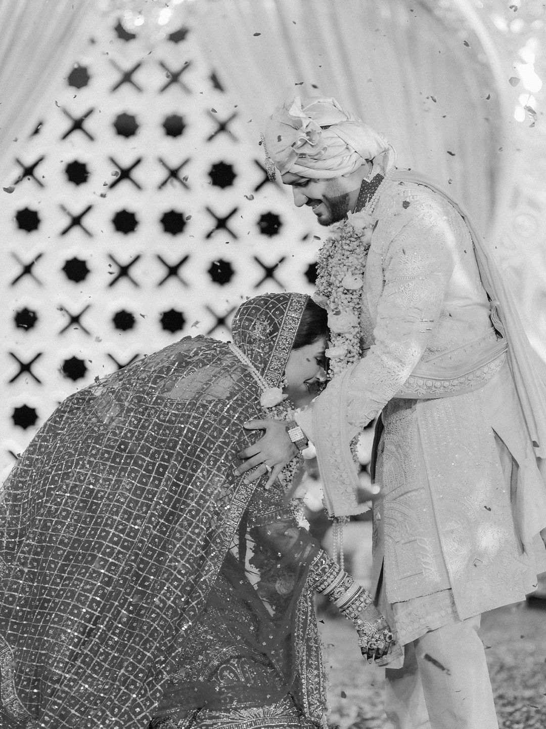 Black and white photo of Indian bride bowing to touch groom's feet during traditional wedding ritual.