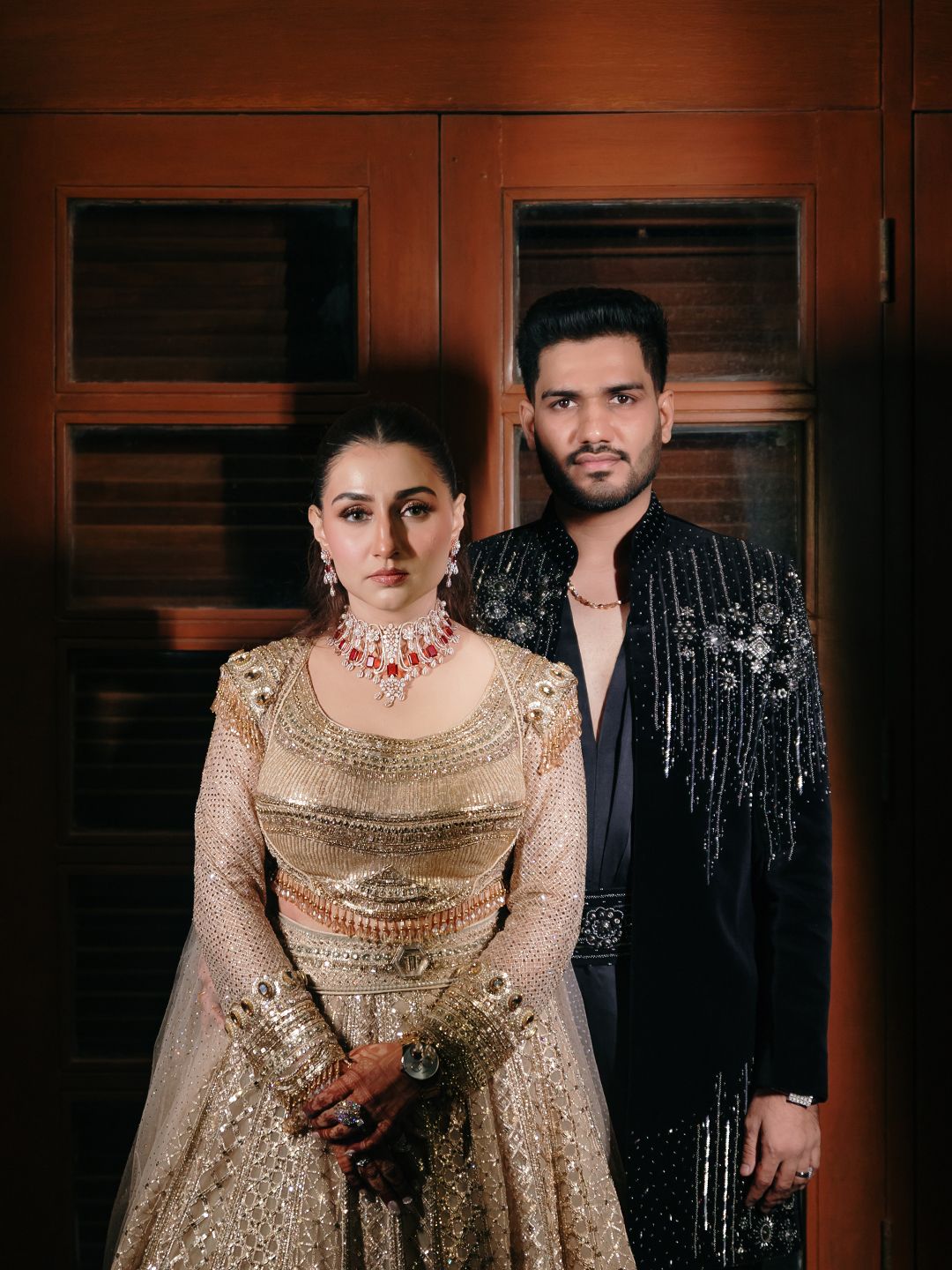 Indian couple in gold embroidered lehenga and black sequined suit with ruby necklace against wooden backdrop.