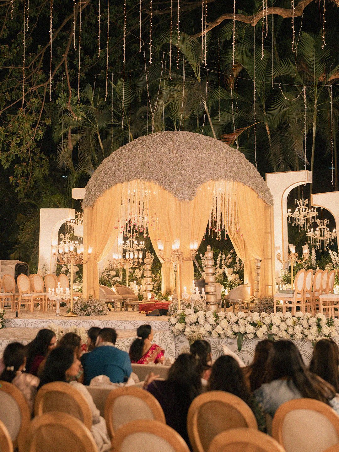 Luxurious outdoor wedding mandap with floral dome, crystal chandeliers, and draped curtains.
