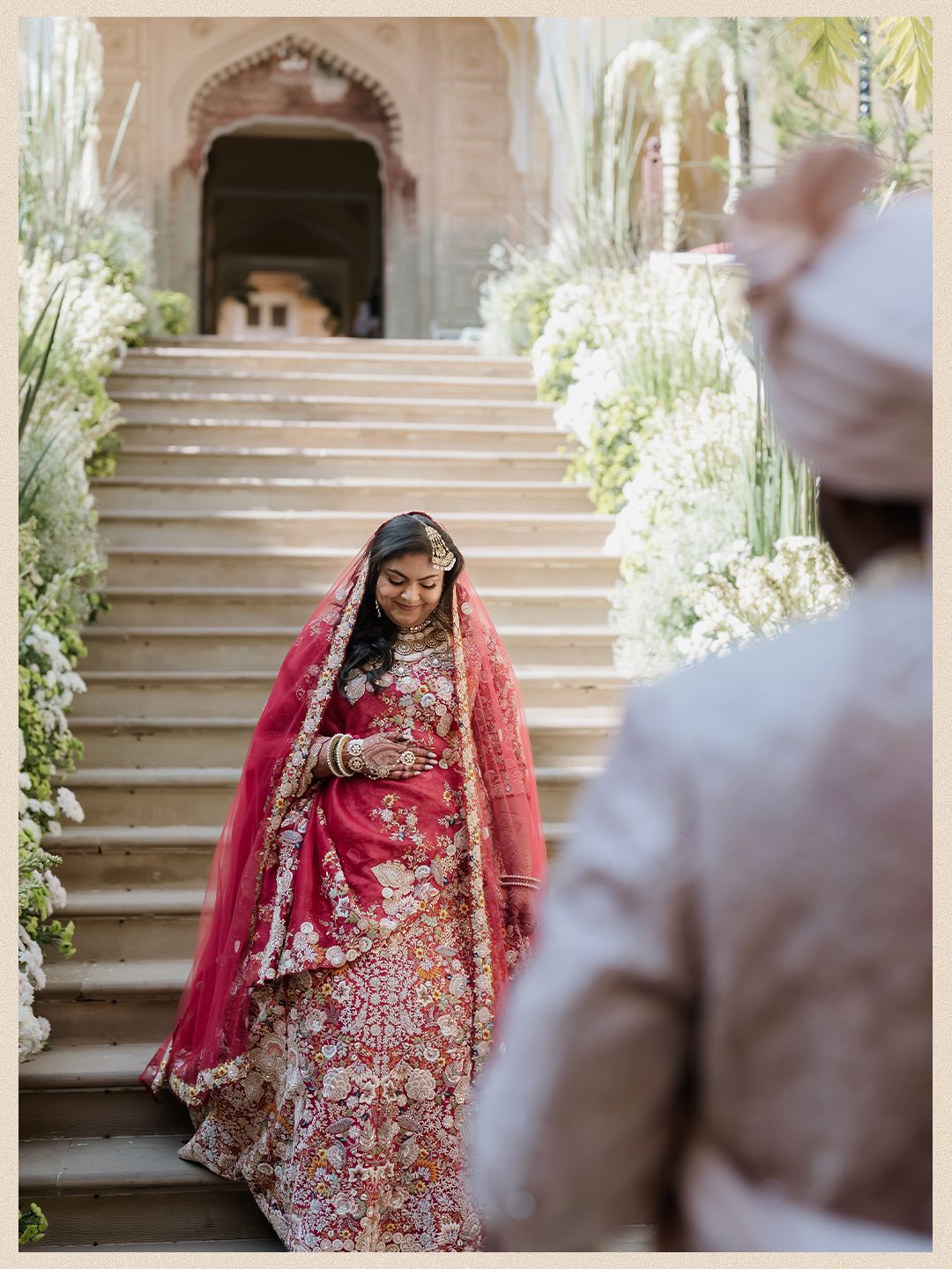 Indian bride descends ornate stone staircase in floral red lehenga with pink dupatta at royal palace wedding venue.