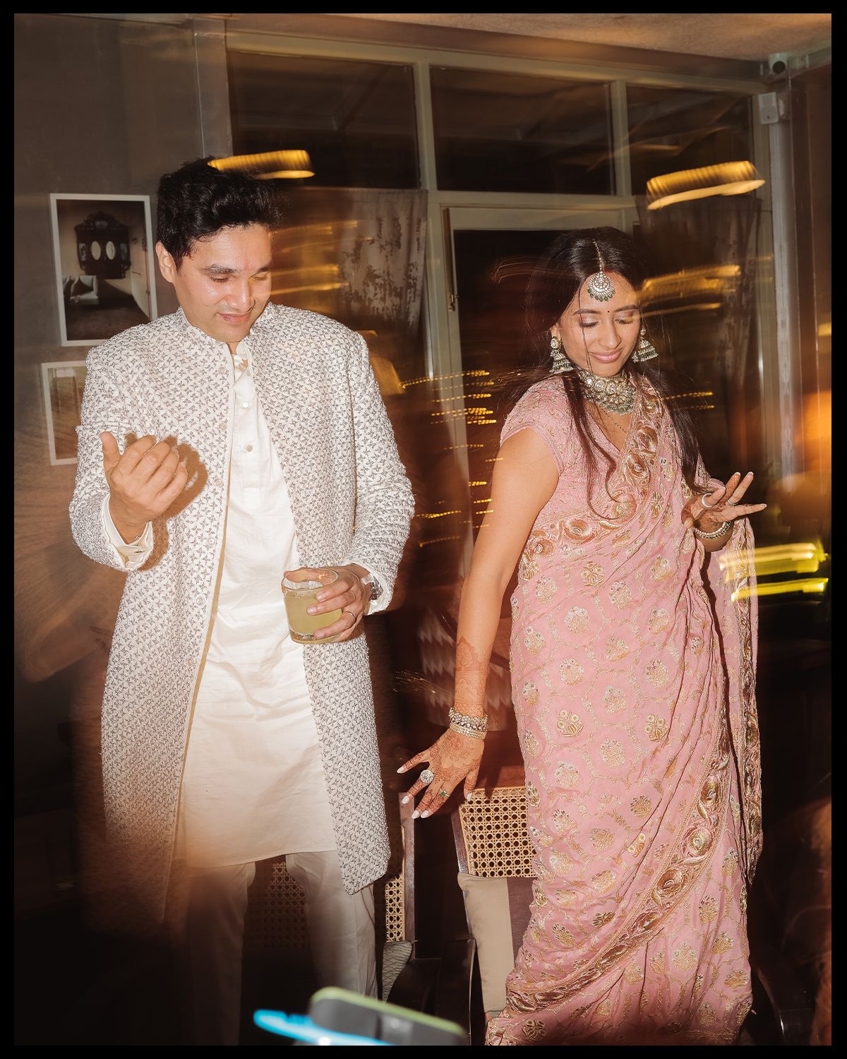 Indian couple dancing at wedding celebration, groom in white sherwani and bride in pink embroidered saree with jewelry.