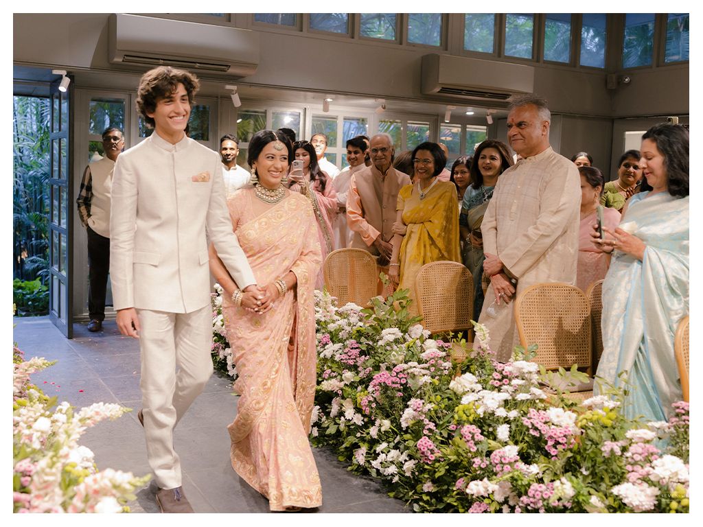 Indian groom in white sherwani walking bride down floral aisle surrounded by wedding guests.
