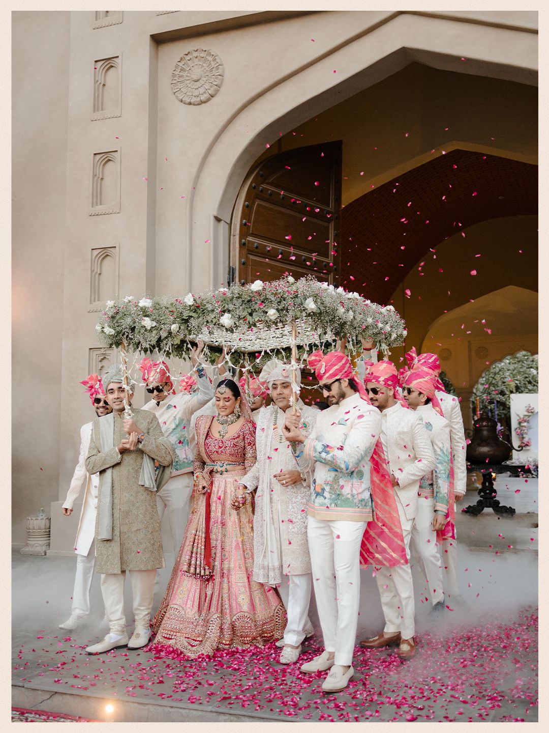 Indian bride and groom in pink and gold wedding attire walking under floral canopy with rose petals falling.