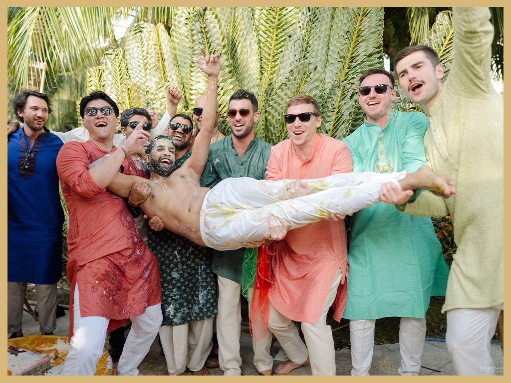 Groomsmen in colorful kurtas joyfully lift groom during fun Indian wedding haldi ceremony outdoors.
