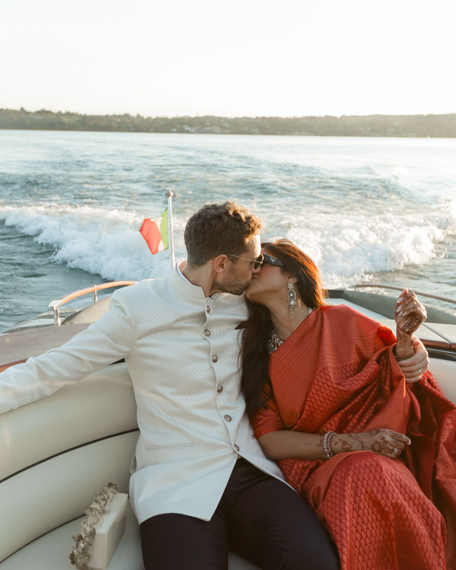 Couple sharing a romantic kiss on a speedboat, bride in red saree and groom in white sherwani.