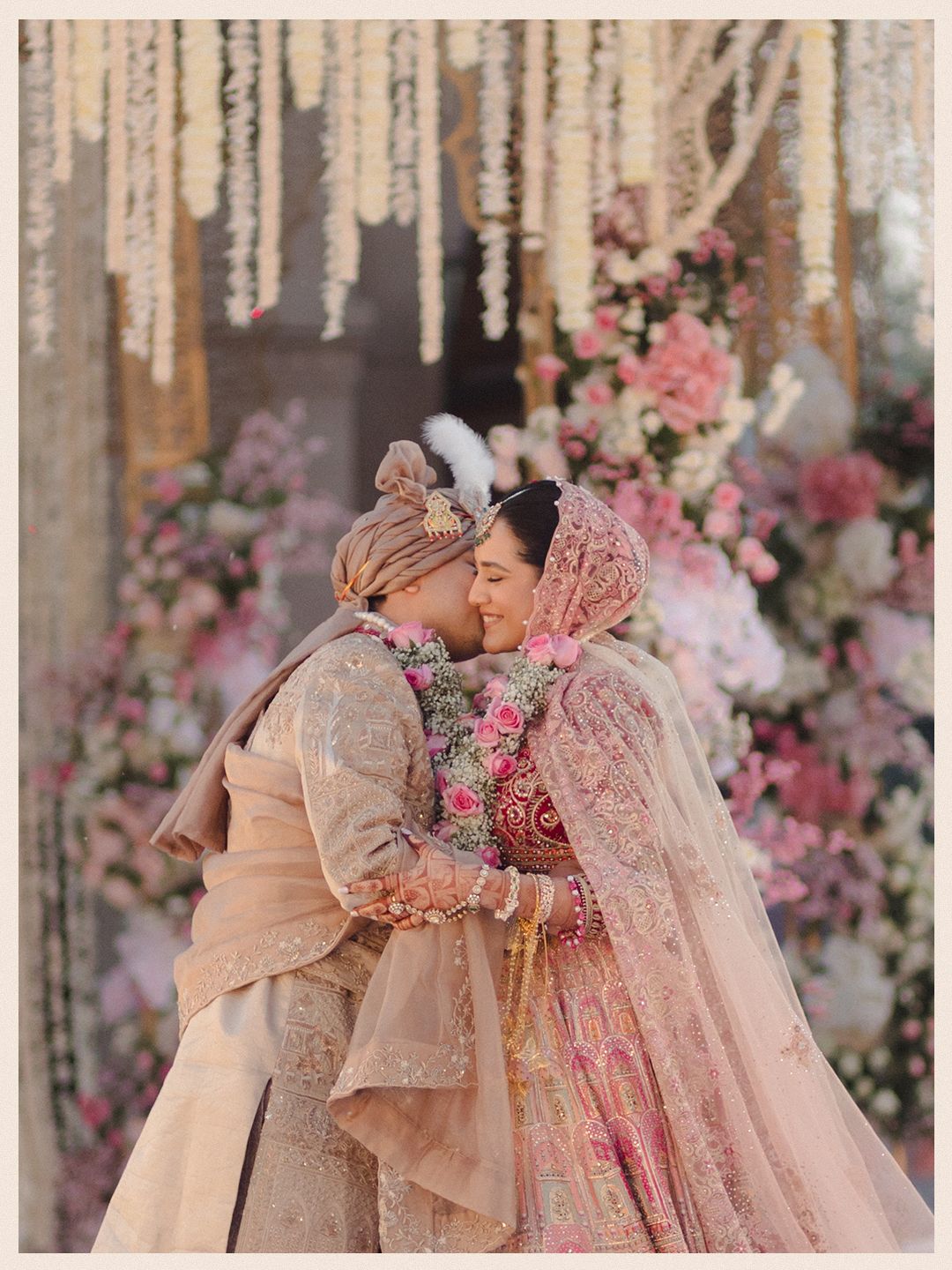 Indian bride and groom sharing a joyful embrace under floral canopy surrounded by pink and white flowers.