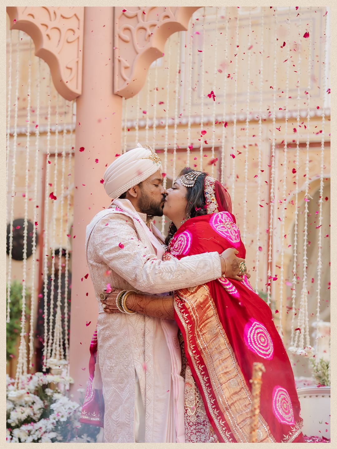 Indian bride and groom share romantic kiss under falling rose petals at traditional wedding ceremony.