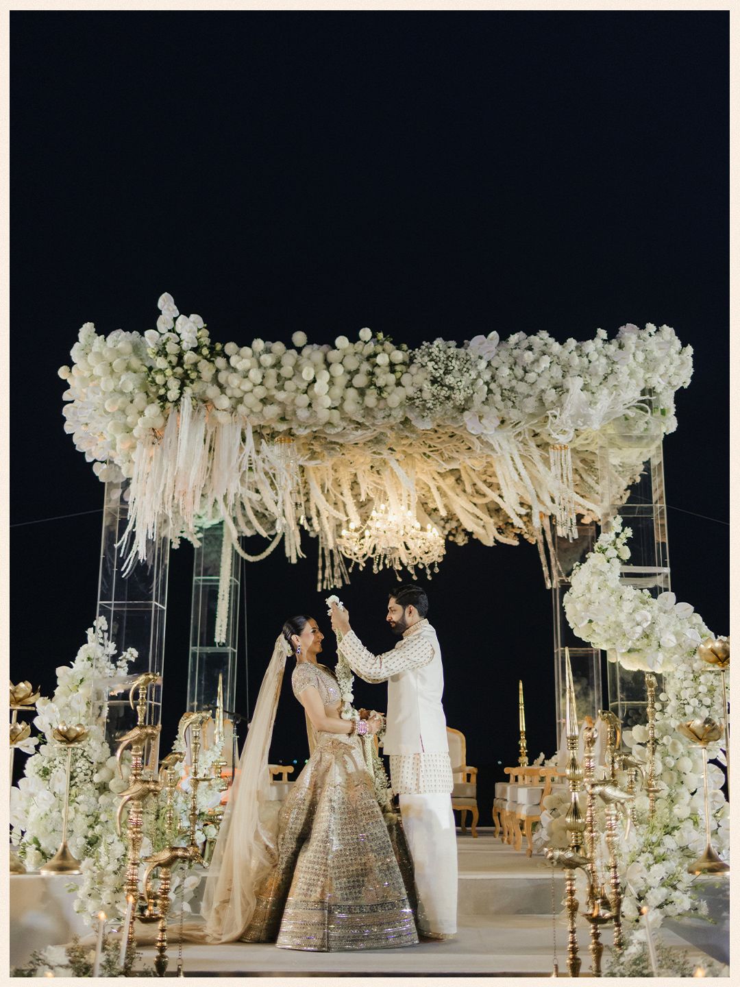 Couple stands under stunning white floral mandap with chandelier at elegant Indian wedding ceremony.