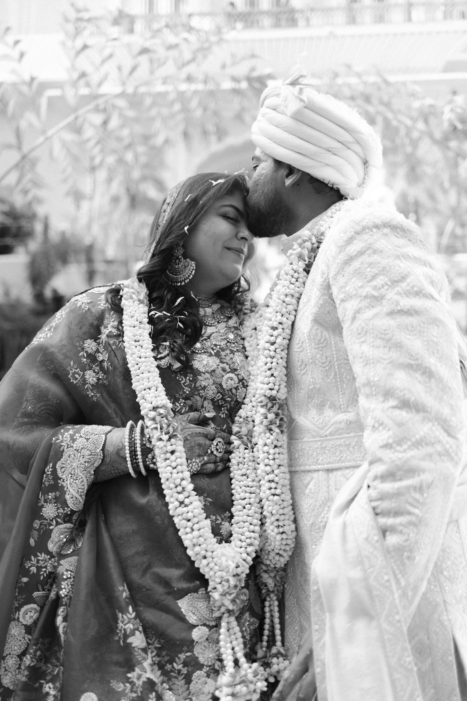 Black and white photo of Indian bride and groom sharing a tender moment wearing floral garlands.