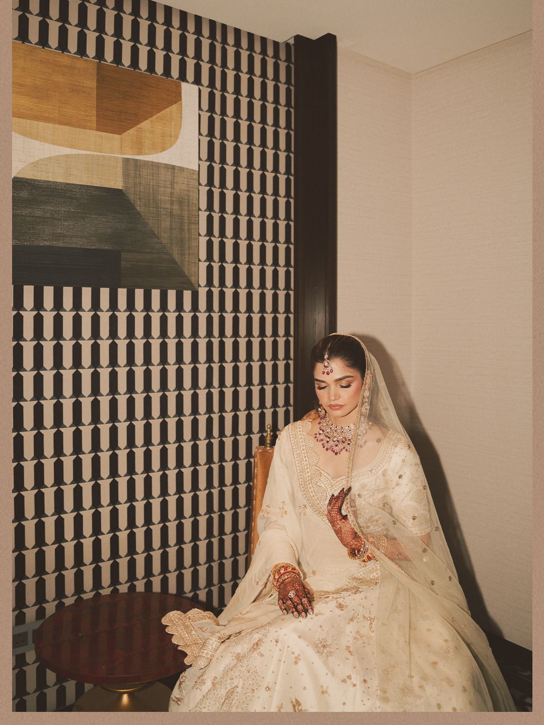Indian bride in cream bridal lehenga with dupatta examining mehndi hands in hotel suite.