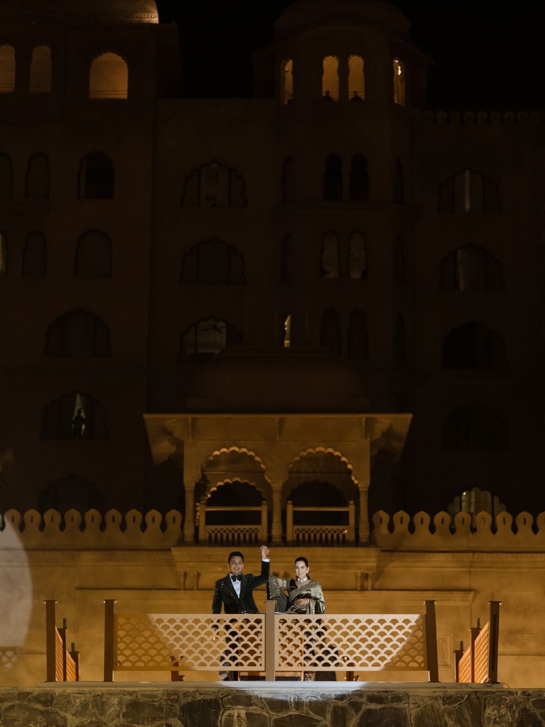 Couple stands on illuminated balcony of a grand Rajasthani palace at night during wedding celebration.