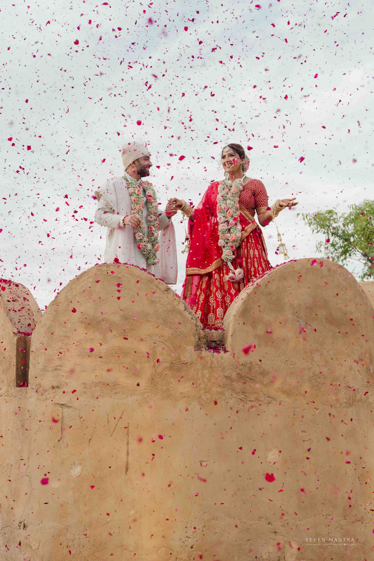 Indian couple in red and white wedding attire celebrating with colorful flower petals at fort.