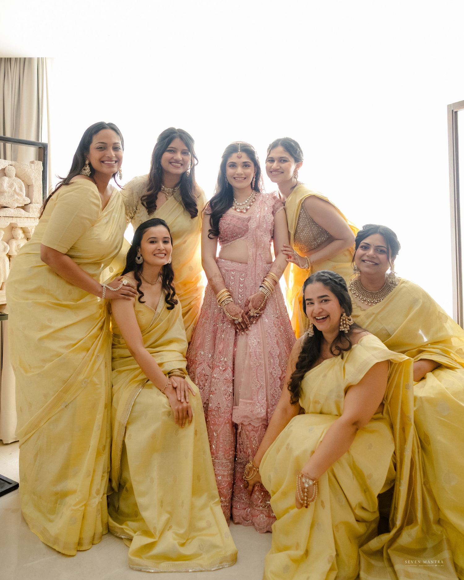 Indian bride in pink lehenga surrounded by bridesmaids in yellow sarees at wedding celebration.