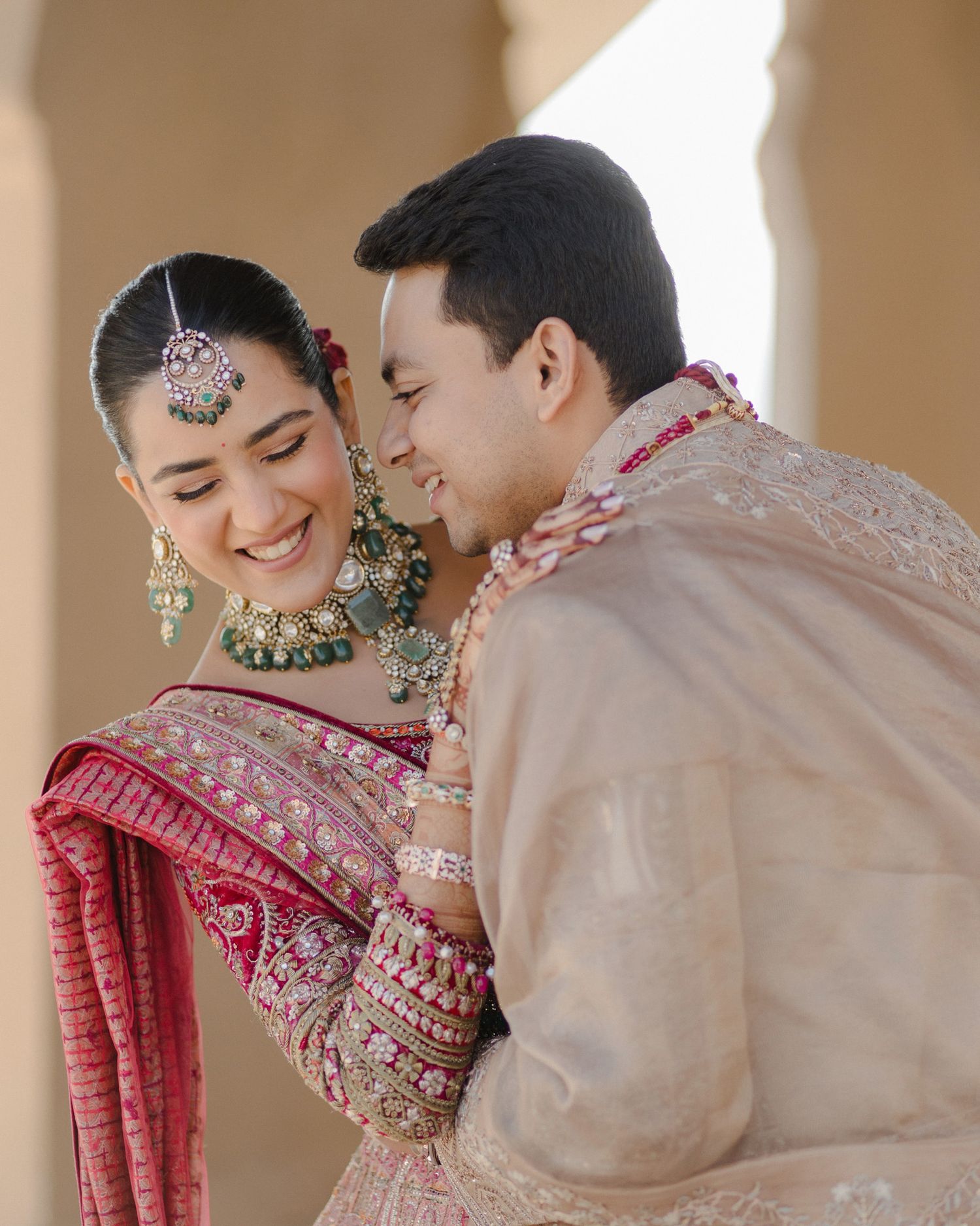 Smiling Indian bride in red lehenga with green jewelry embraced by groom in beige sherwani.
