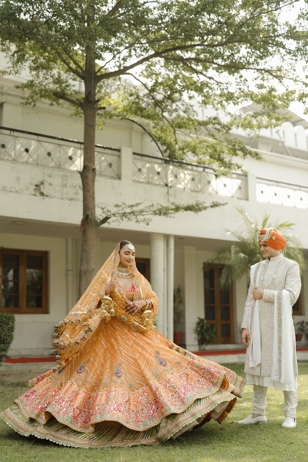 Bride in orange embroidered lehenga twirls joyfully as groom in ivory sherwani watches outdoors.