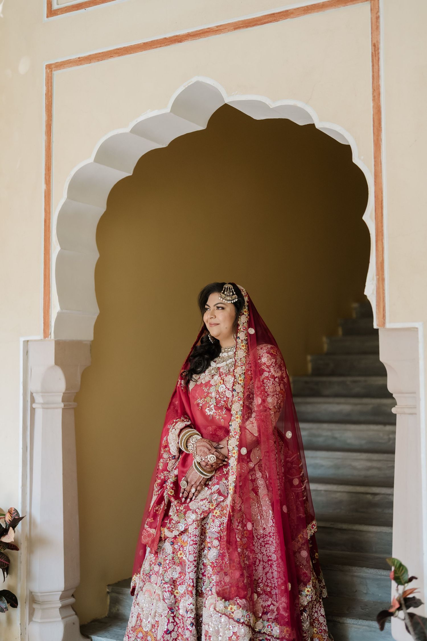 Indian bride in stunning red and gold embroidered lehenga standing in an ornate white arched doorway.
