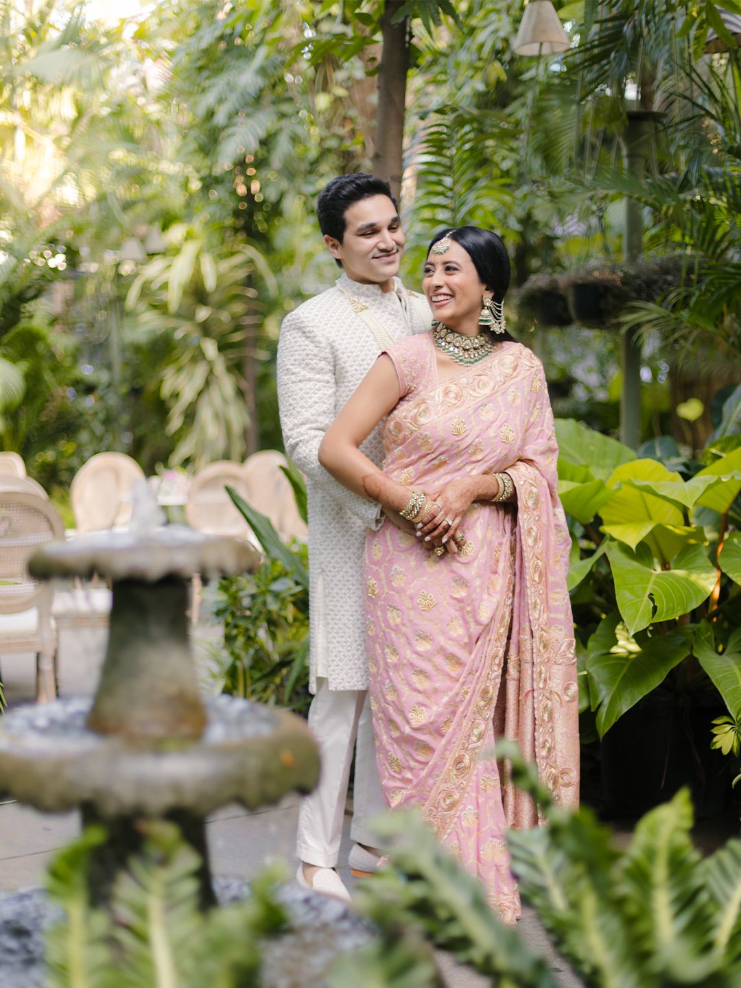 Couple poses together in lush green garden, groom in white sherwani and bride in pink embroidered saree.