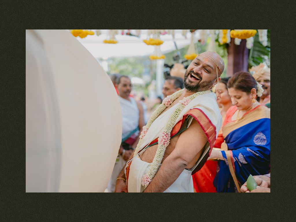 Bald groom laughs joyfully during a vibrant South Indian wedding ceremony surrounded by guests in colorful attire.