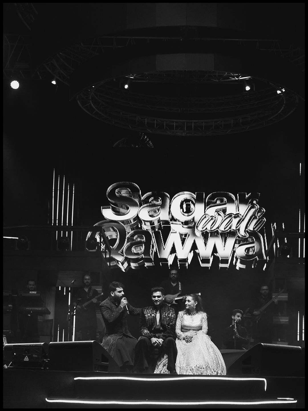 Black and white photo of couple seated on stage beneath illuminated Sagar wali Qawwali sign.