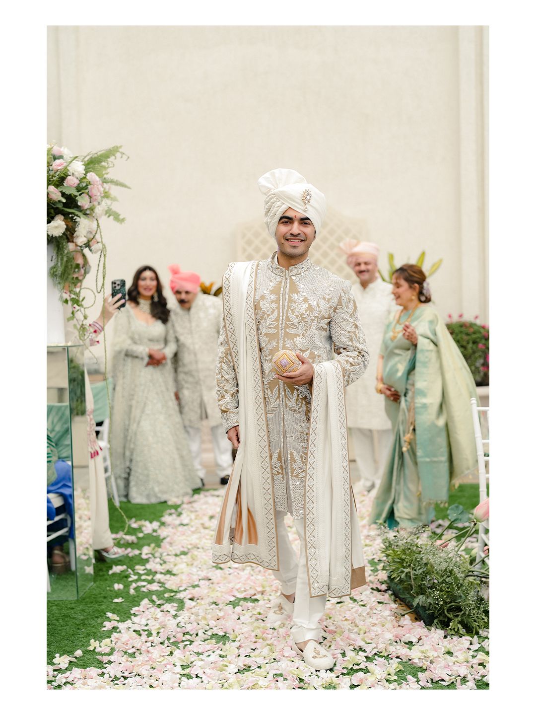 Handsome Indian groom in ivory embroidered sherwani and white turban walking down a rose petal aisle.