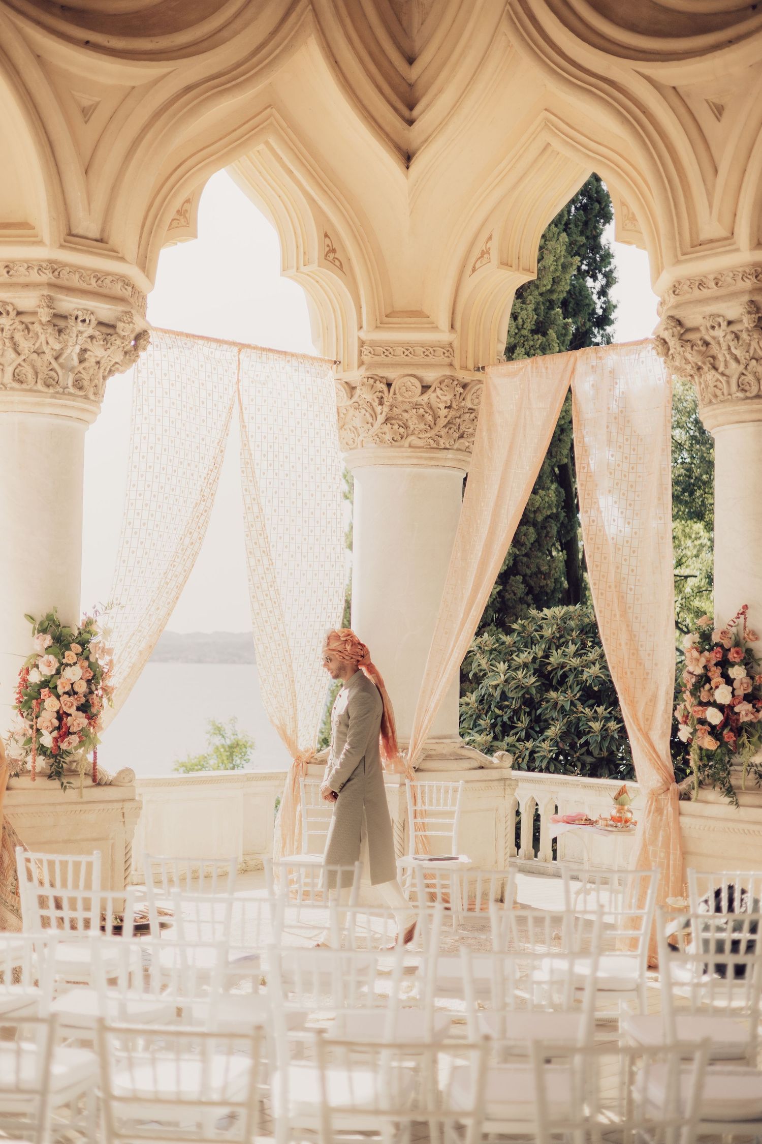 Wedding mandap setup with ivory drapes and pink florals in grand arched hall overlooking the sea.