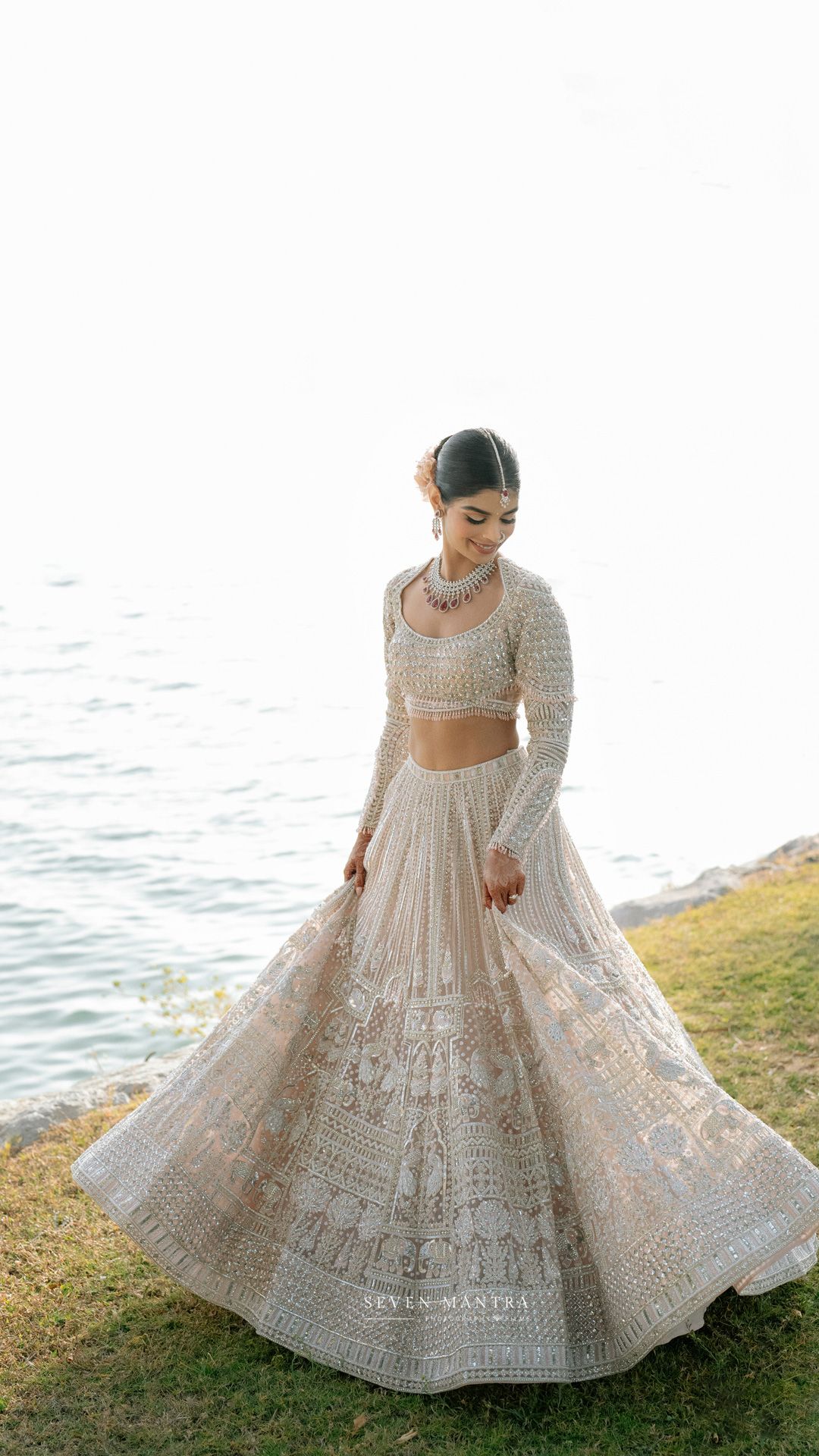 Bride twirling ivory embroidered lehenga with intricate motifs near a scenic waterfront location.