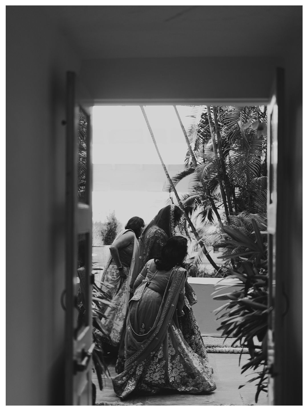 Black and white photo of women in sarees viewed through doorway with tropical garden backdrop.