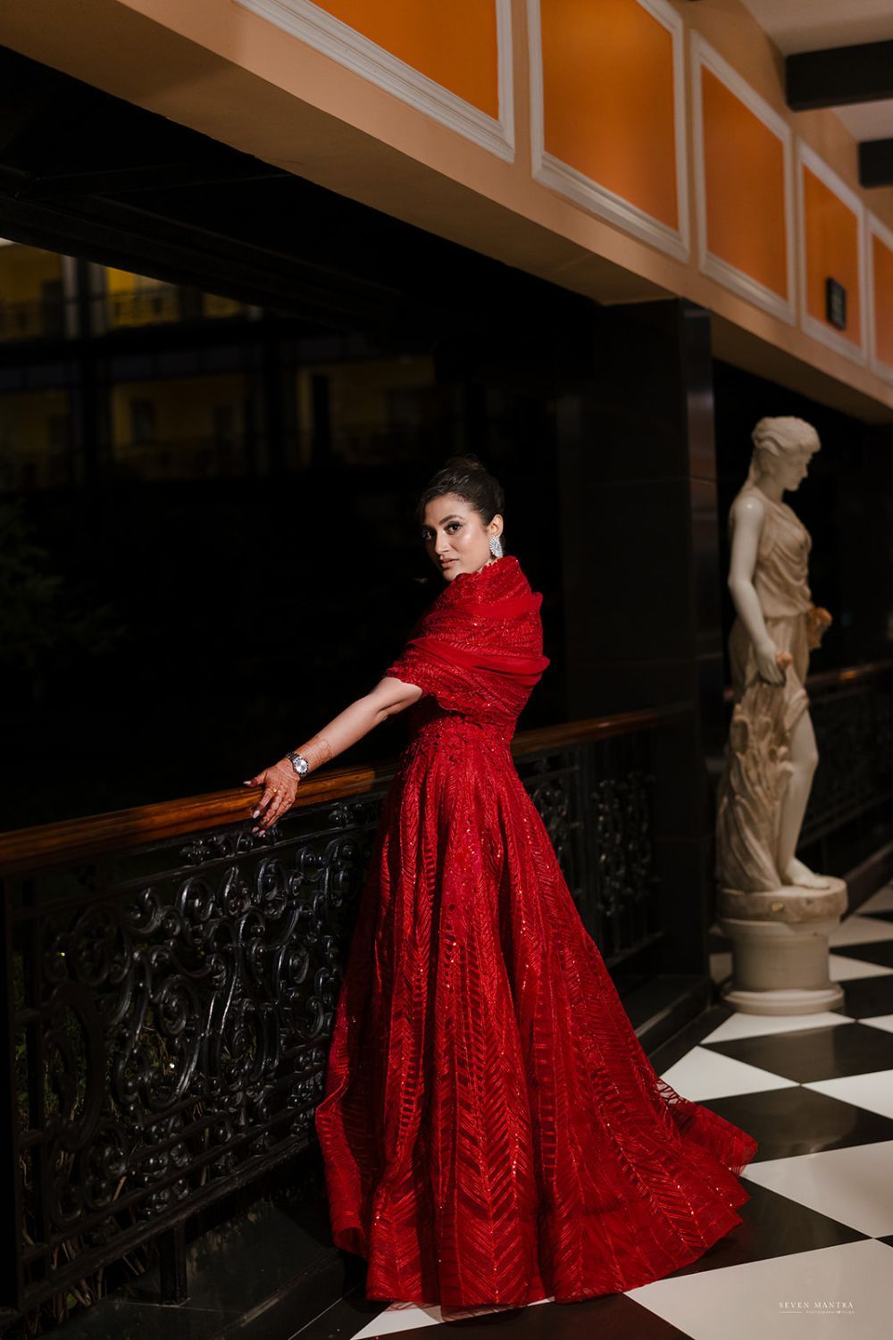 Elegant woman in red embellished ball gown poses on ornate staircase beside classical sculpture.