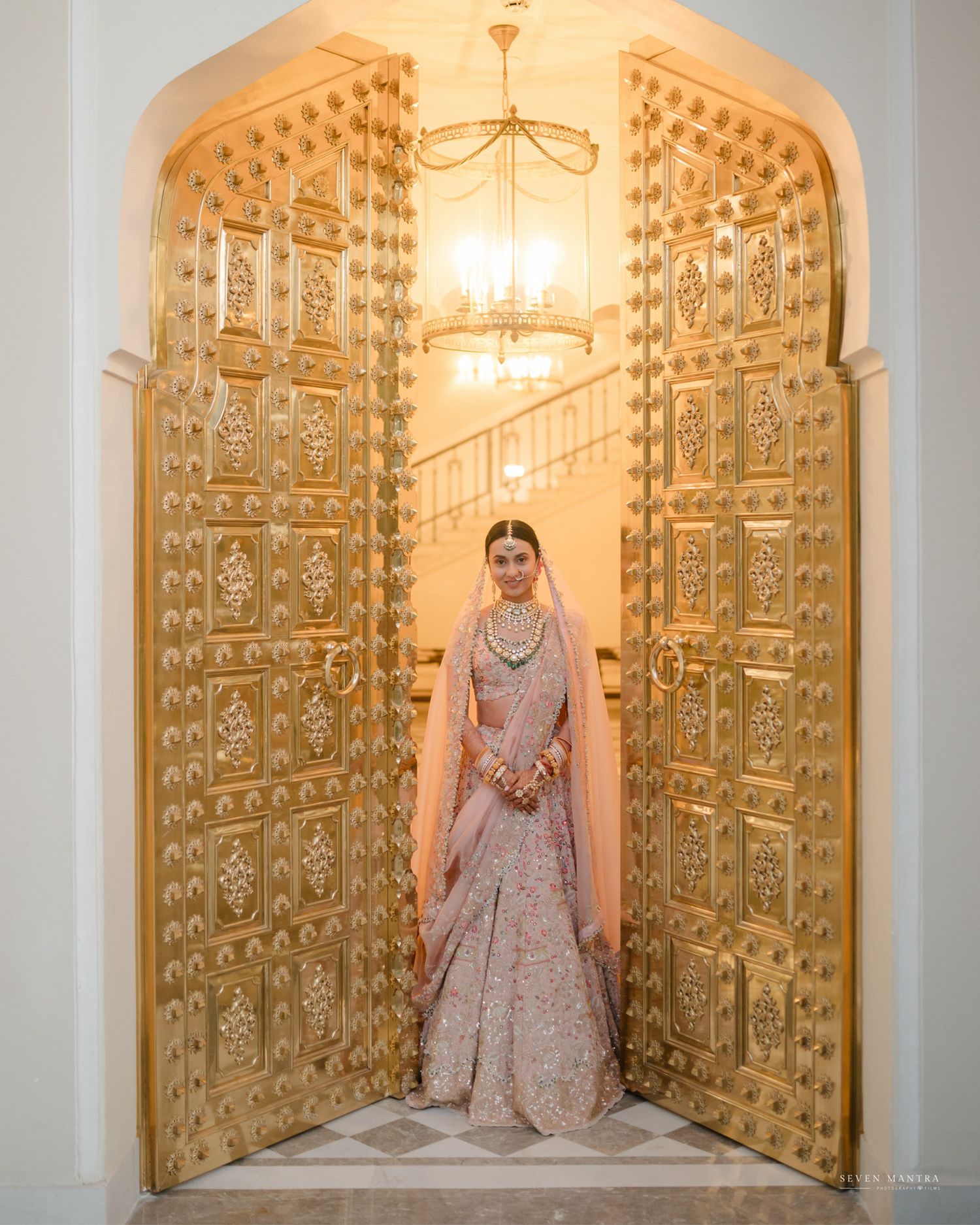 Indian bride in silver lehenga posing between ornate golden doors with chandelier backdrop at wedding venue.