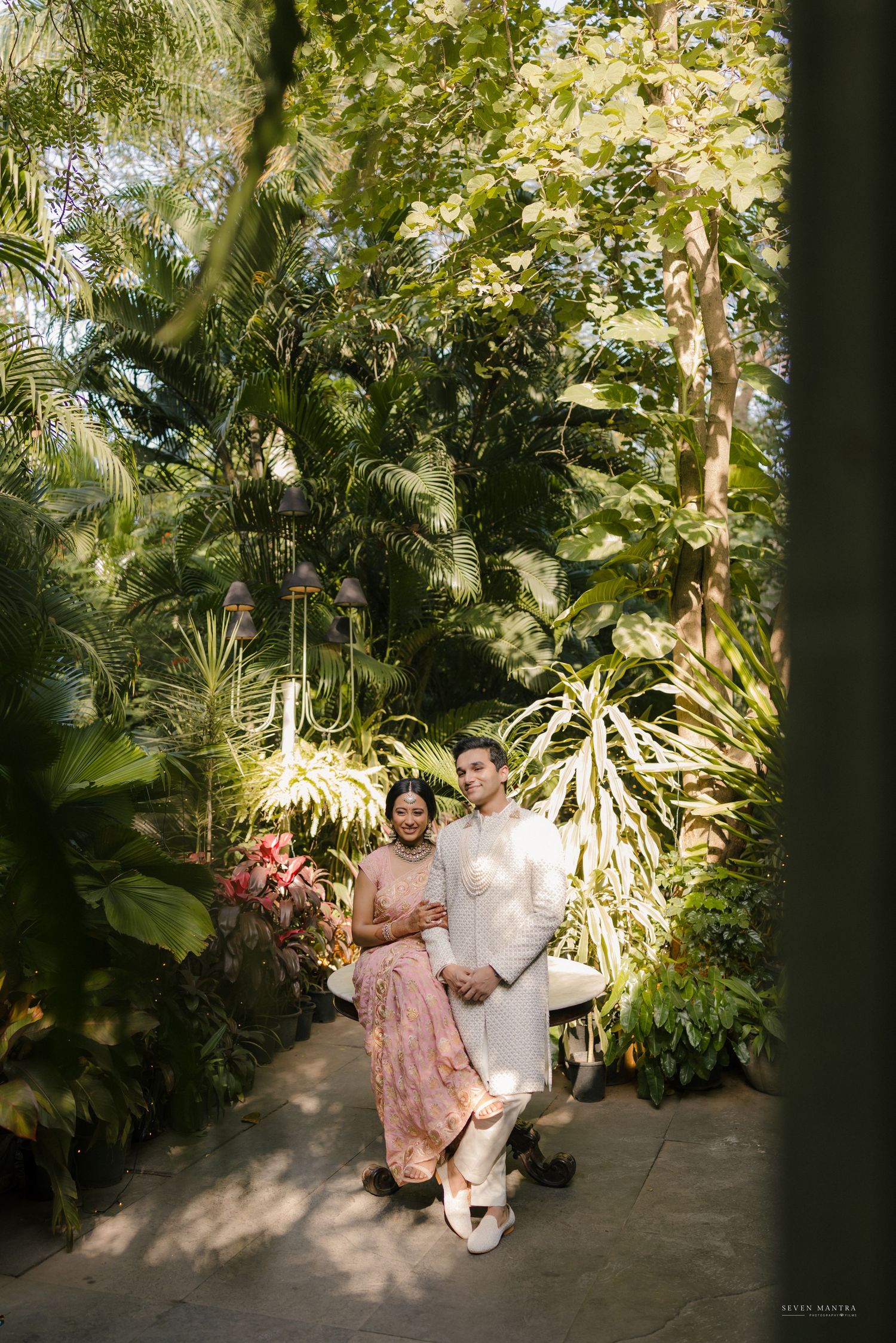 Elegant couple in white and pink Indian wedding attire posing in lush tropical garden.