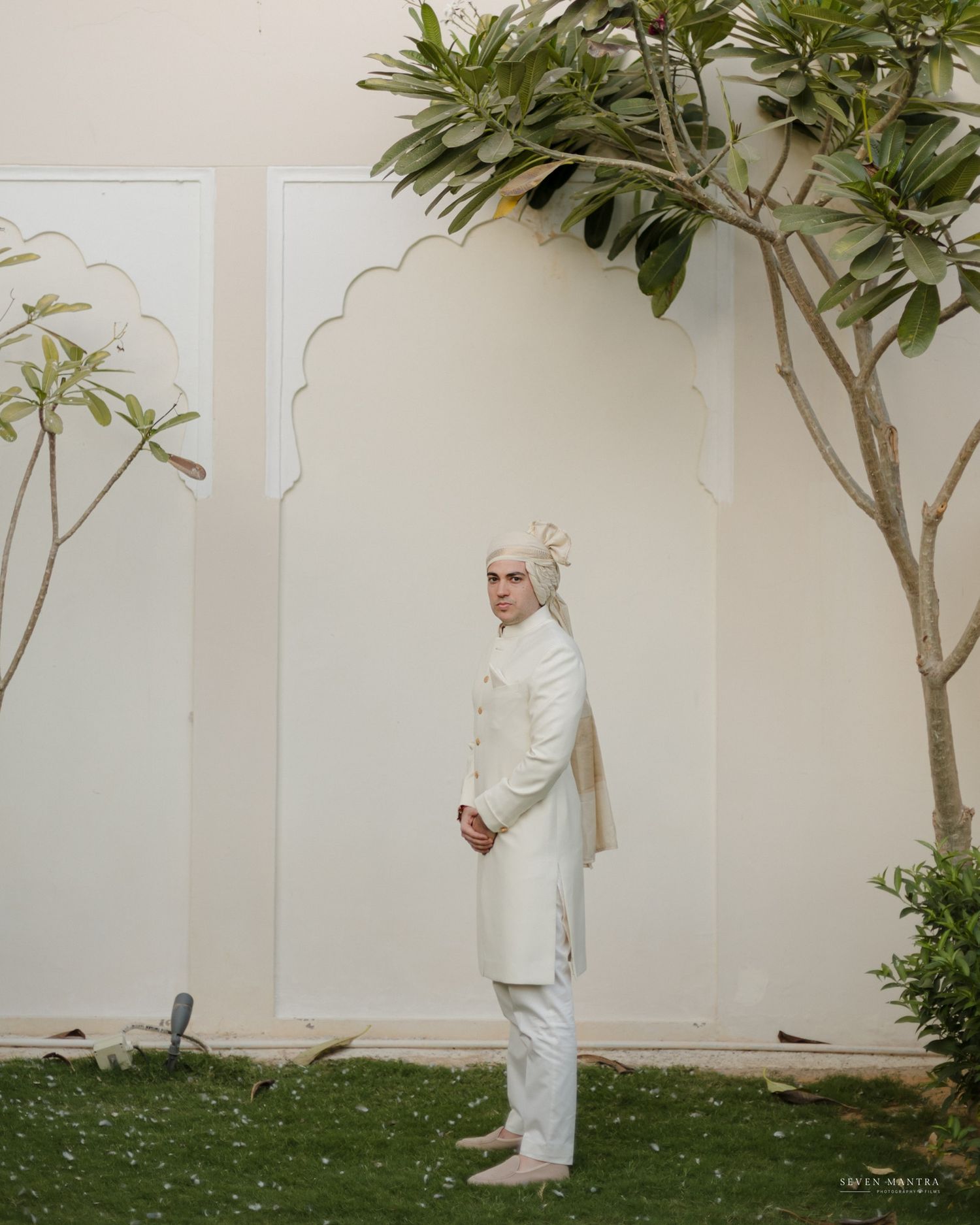 Groom in elegant white sherwani and turban standing in an arched garden courtyard on wedding day.