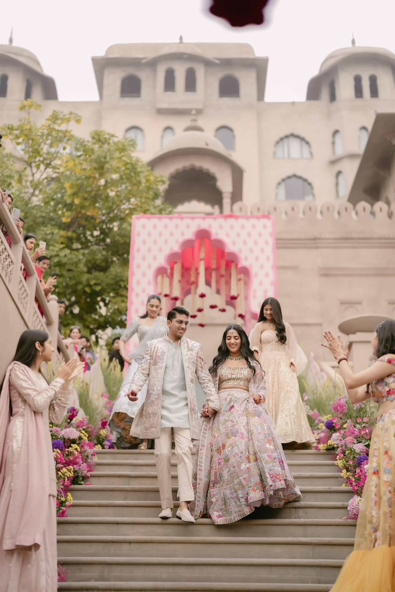 Indian bride and groom descending grand staircase in colorful lehenga at palace wedding venue.
