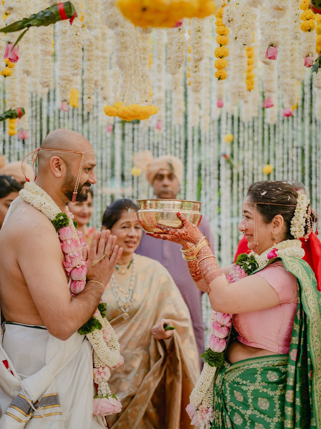 Indian bride offers ritual bowl to groom under floral mandap during traditional wedding ceremony.