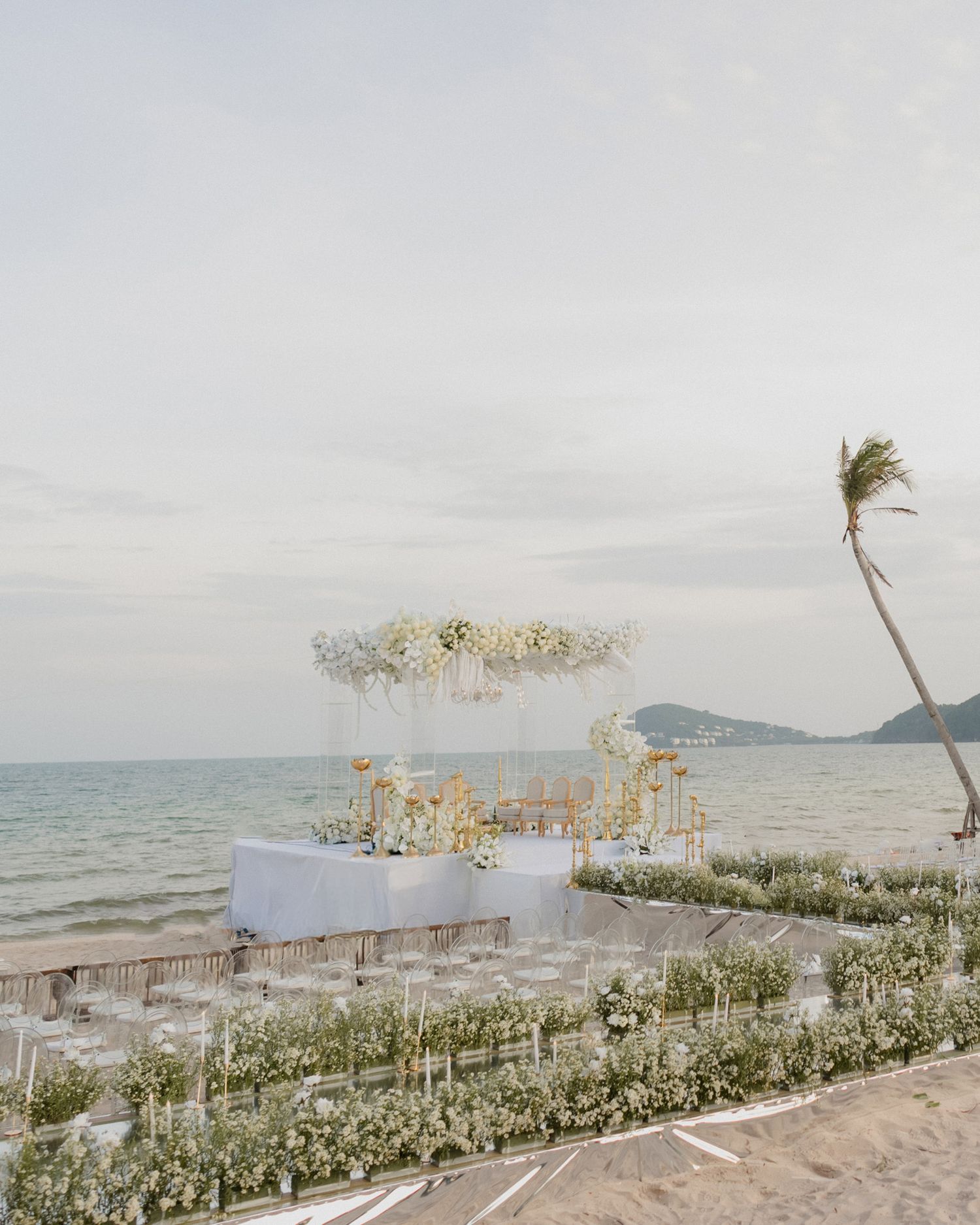 Elegant beachside wedding ceremony setup with white floral arch, chairs, and ocean backdrop at sunset.