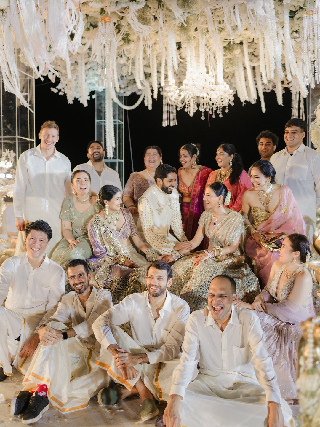Joyful wedding group photo with bride, groom, and guests under white floral chandelier decor.