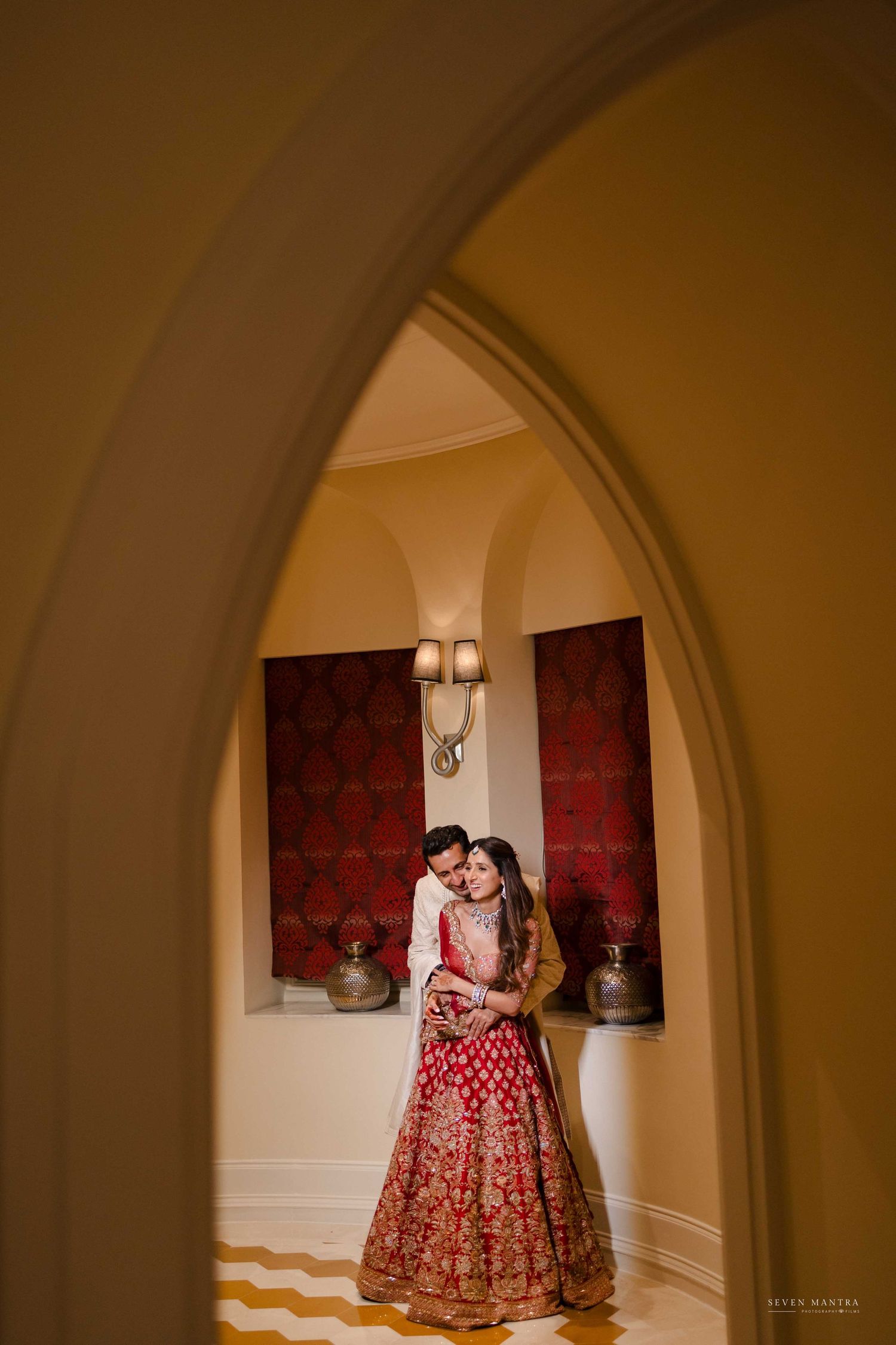 Bride in red embroidered lehenga and groom embrace framed by ornate archway in golden corridor.