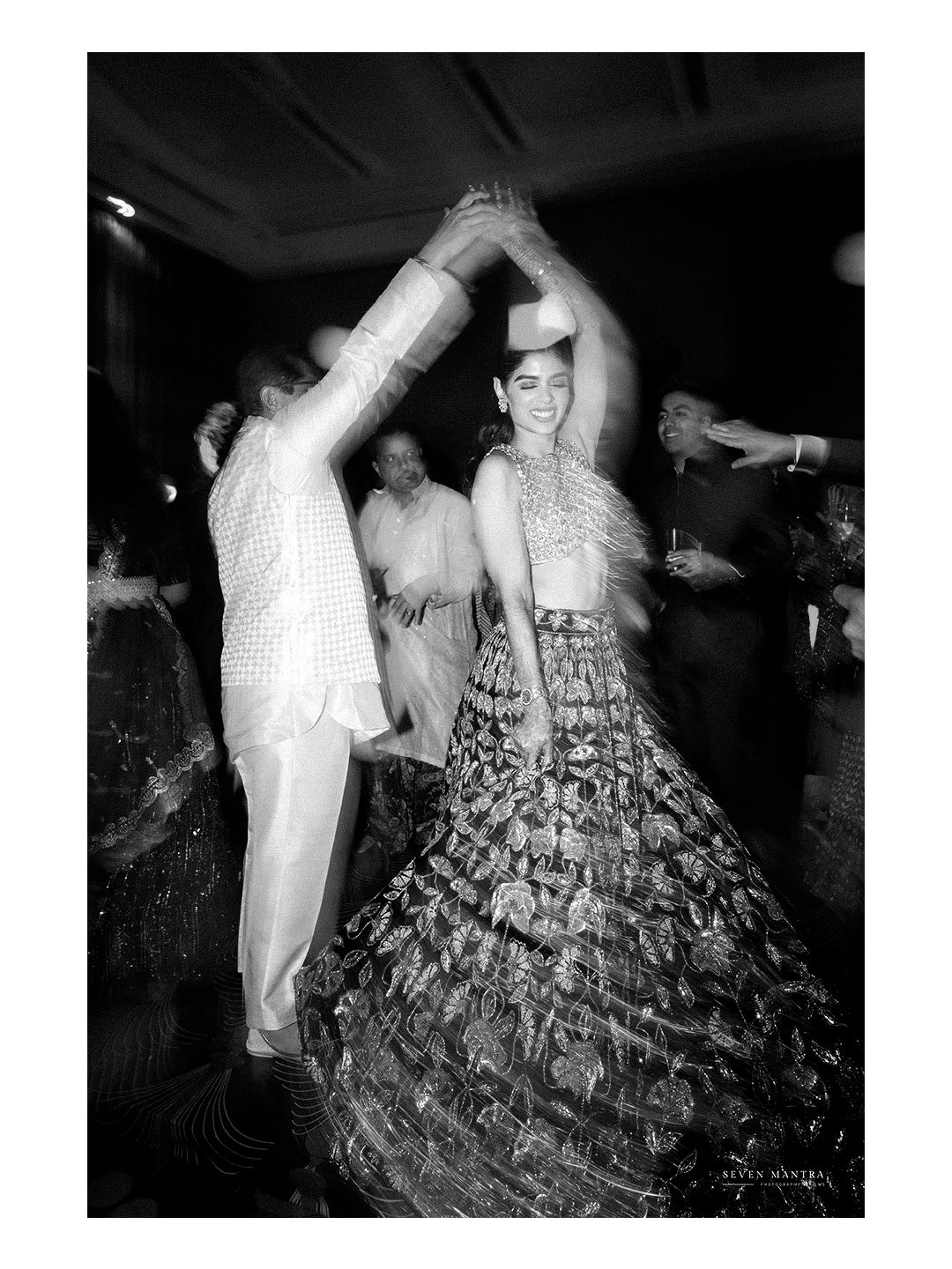 Black and white photo of Indian bride in floral lehenga dancing joyfully at wedding reception.