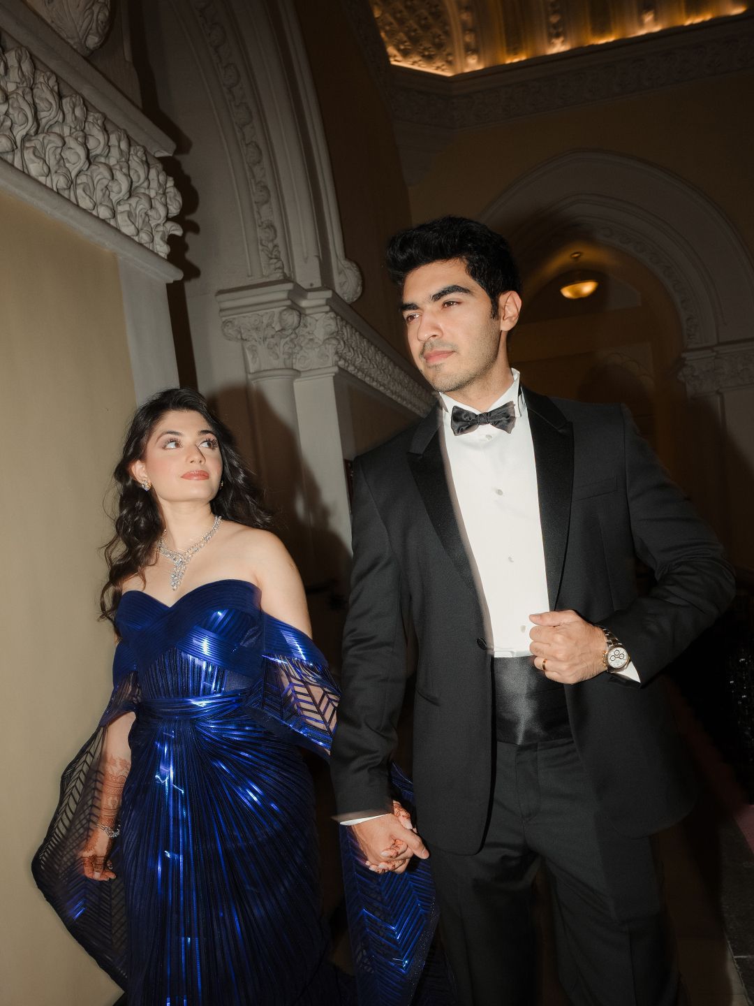 Bride in metallic blue gown holds hands with groom in black tuxedo at ornate wedding venue.