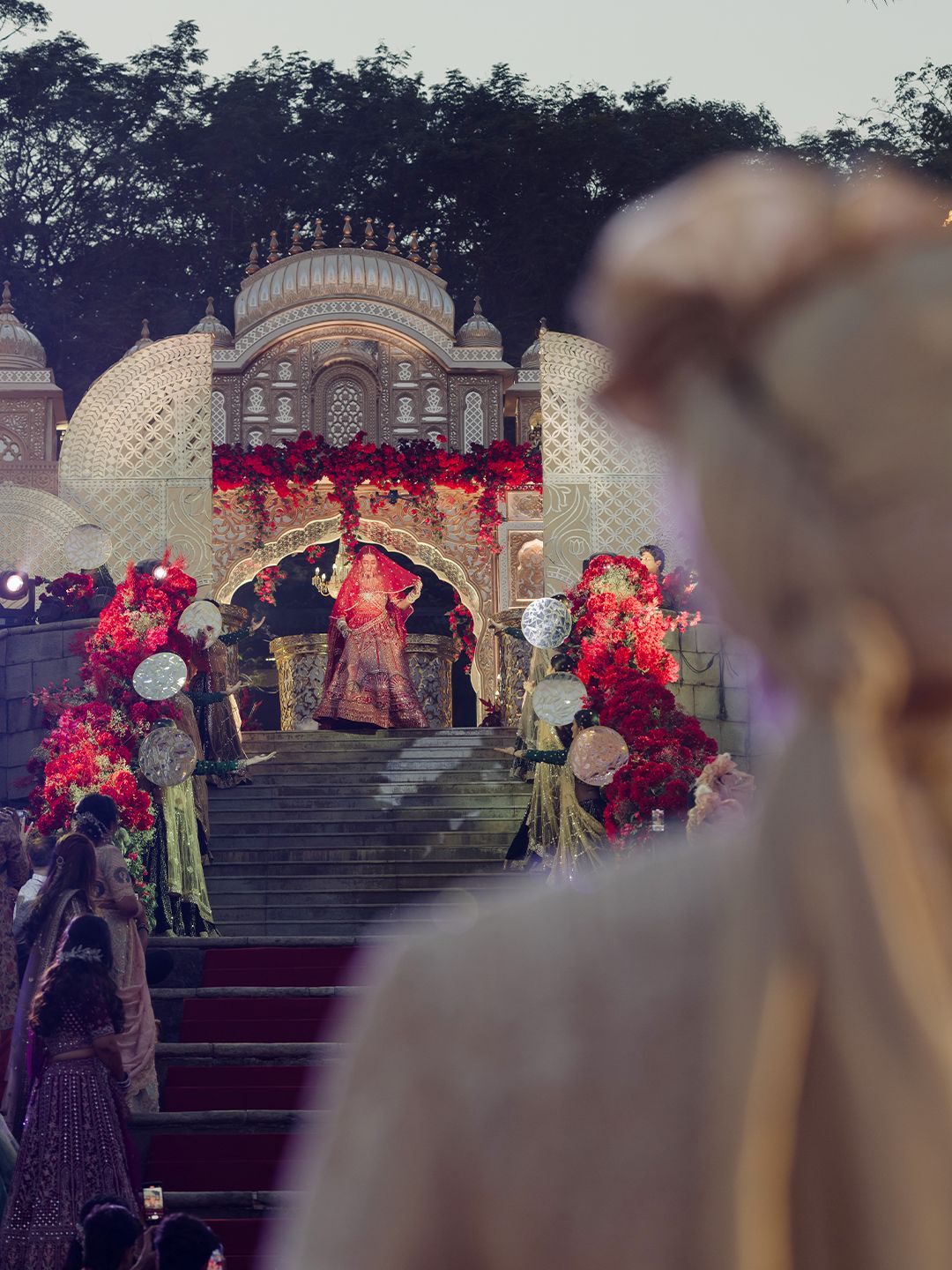 Bride descending grand Mughal-inspired staircase adorned with red floral wedding decorations.