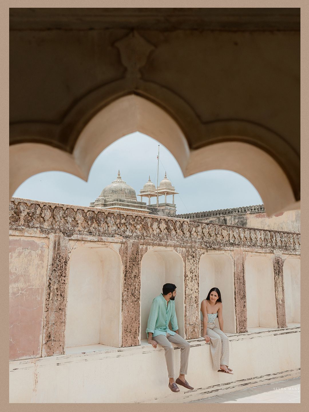 Couple sitting together framed by ornate Mughal arch with palace architecture in background.