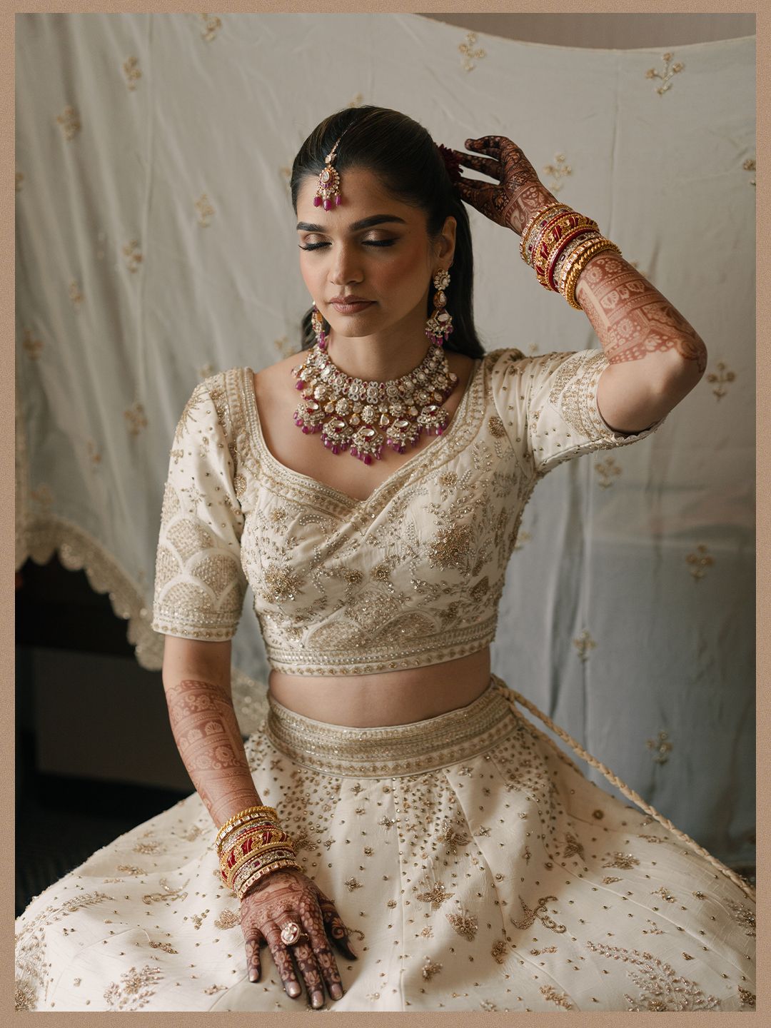 Indian bride in ivory embroidered lehenga with gold jewelry and mehndi hands in natural light portrait.