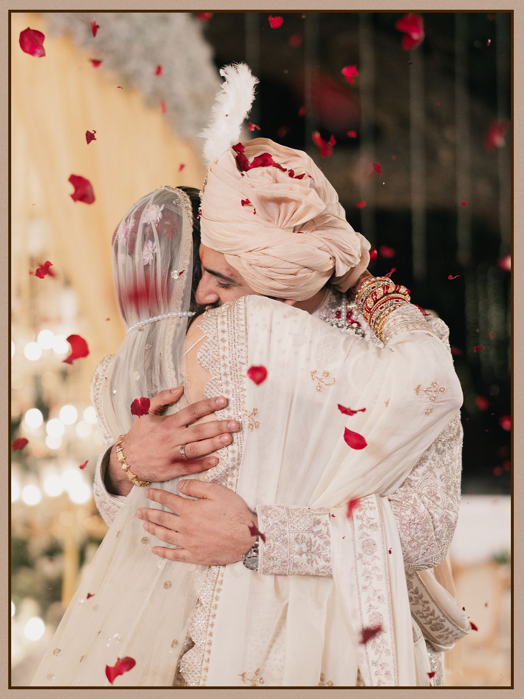 Indian newlyweds in matching ivory outfits sharing emotional embrace under falling rose petals.