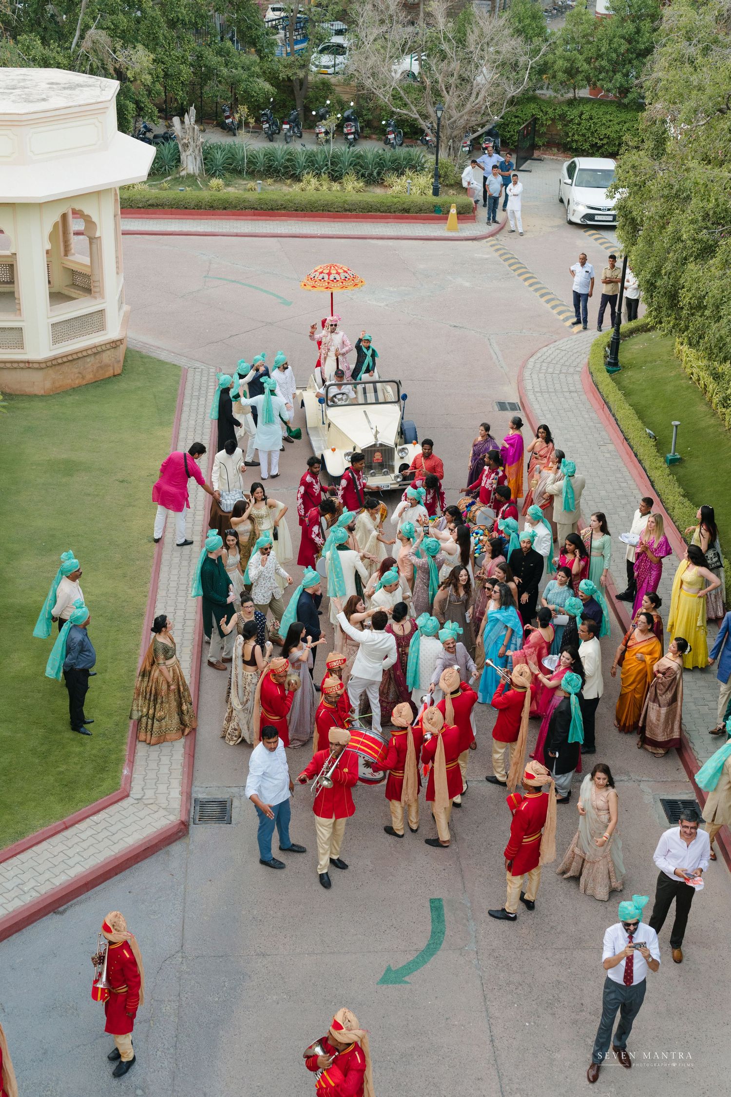 Aerial view of Indian wedding baraat procession with groom on vintage car surrounded by dancing guests.