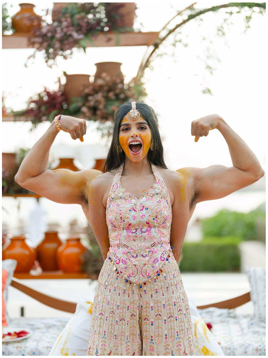 Bride flexes arms playfully with turmeric on face during Indian Haldi celebration.
