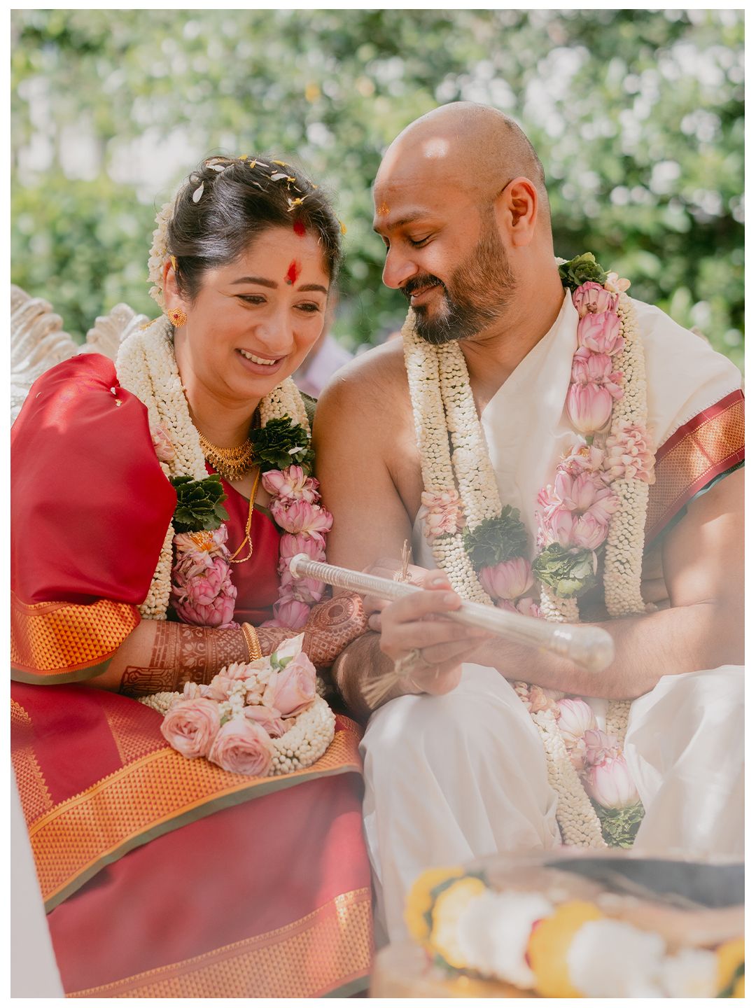 Bride in red saree and bald groom share a joyful moment reading during a South Indian wedding ceremony.