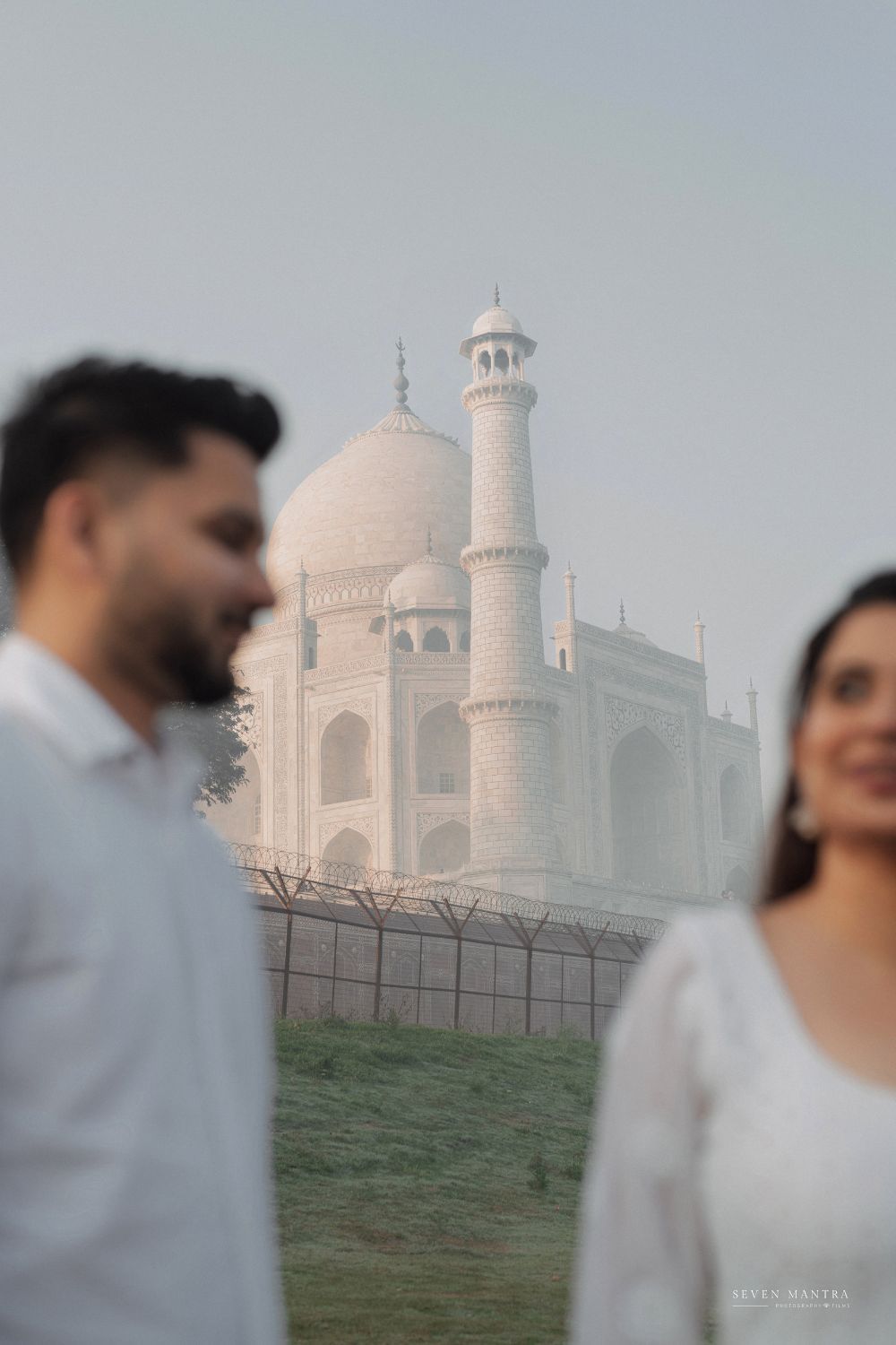 Couple in white outfits stands before the iconic misty Taj Mahal in Agra, India.