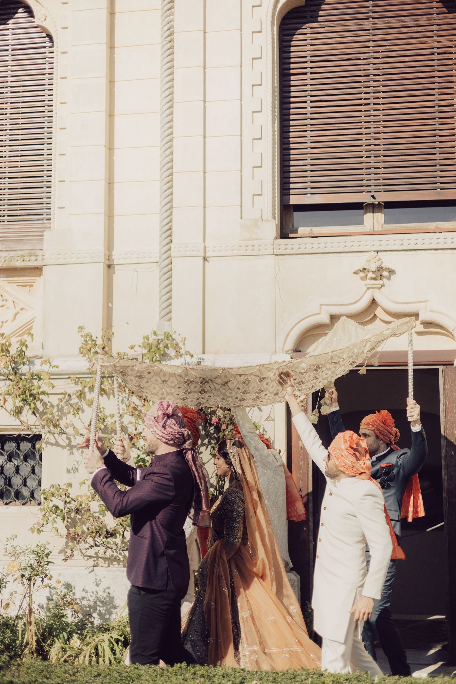 Indian wedding ceremony with guests holding a decorative canopy over the bride and groom outside an ornate building.